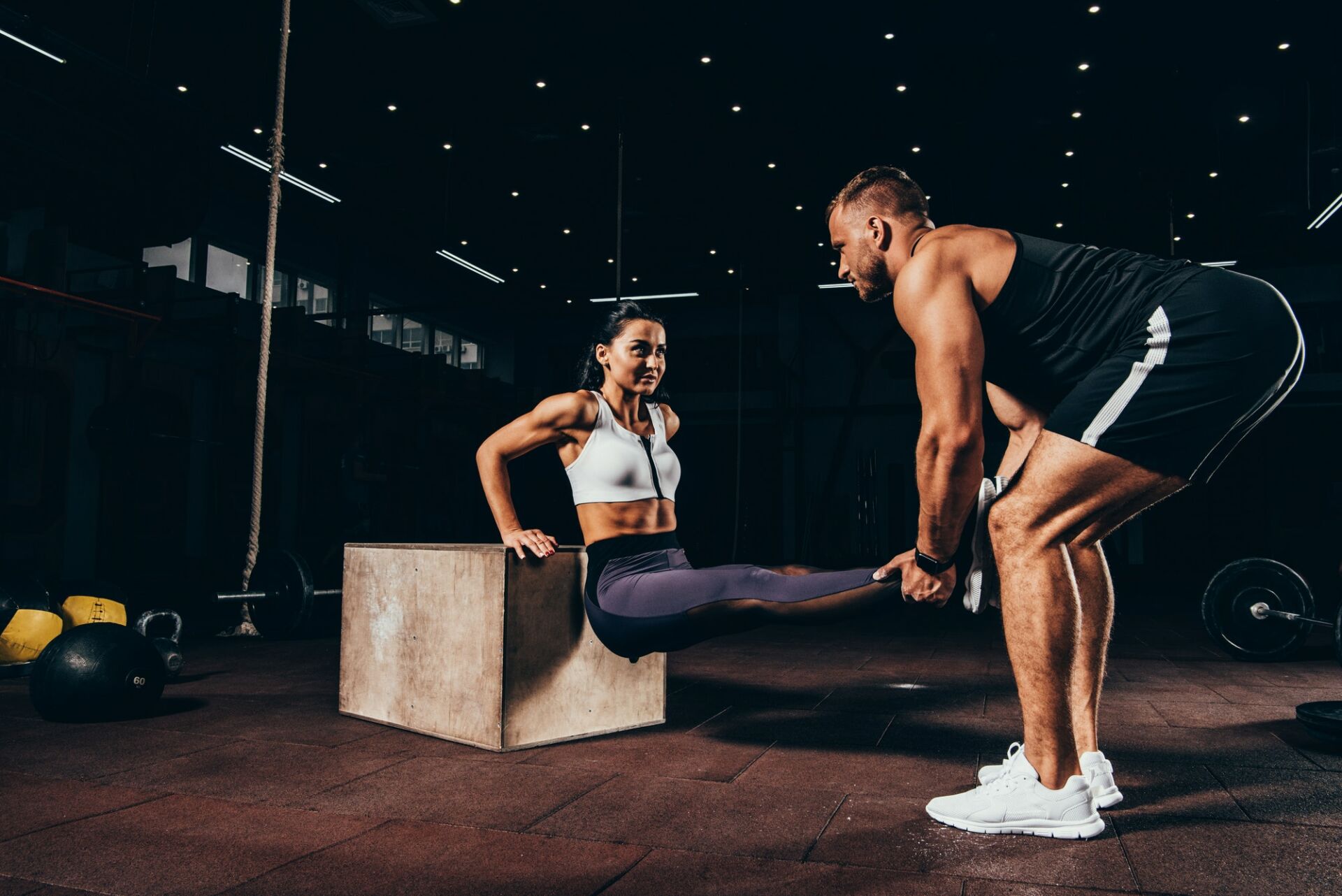 athletic woman exercising with trainer on cube in dark gym