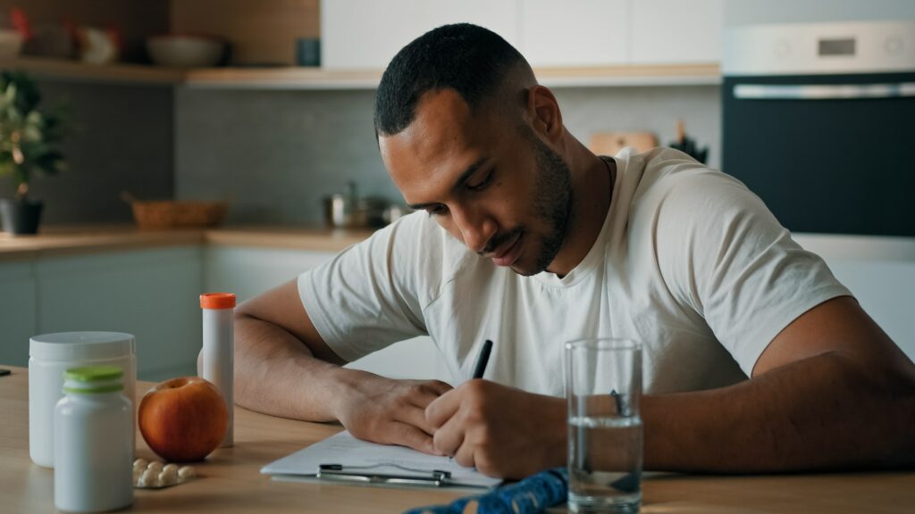 African American male nutritionist man sport fitness yoga trainer sitting in kitchen writing