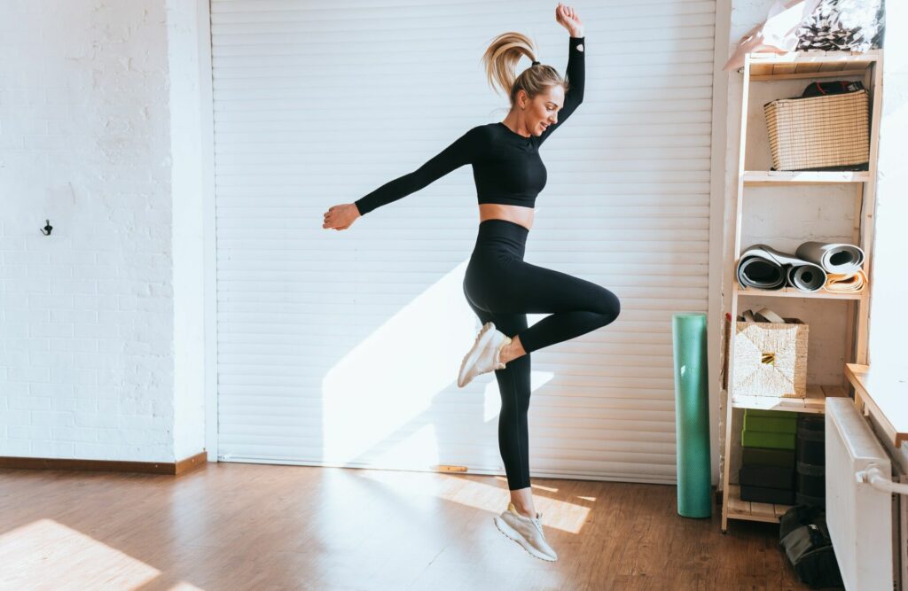 Fit caucasian young blonde woman in black sportswear jumps, training at fitness club on sunny day.